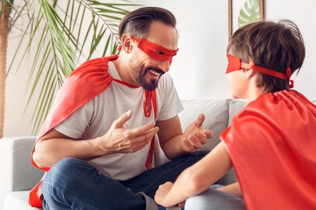 Father and son in superheroe costumes at home sitting on sofa talking laughing cheerful boy back close-upの写真素材