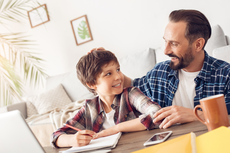 Father and son at home sitting at table dad praising boy for good grades proud while doing homeworkの写真素材