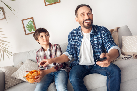 Father and little son at home sitting on sofa boy holding potato chip looking confused at dad watching football eating snack smiling excitedの写真素材