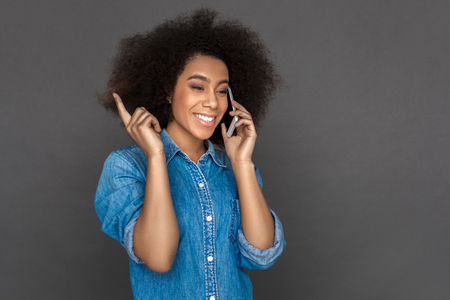 Freestyle. Mulatto woman standing isolated on grey answering phone call smiling cheerfulの写真素材