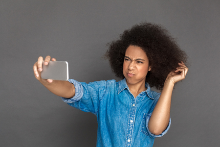 Freestyle. Mulatto woman standing isolated on grey taking selfie on smartphone pouting lips dissatisfied to cameraの写真素材