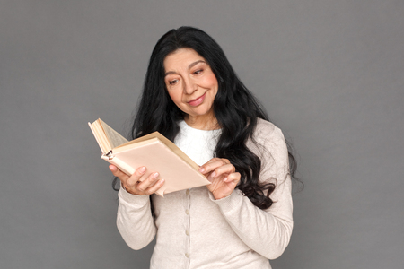 Freestyle. Mature lady standing isolated on grey reading book curiousの写真素材