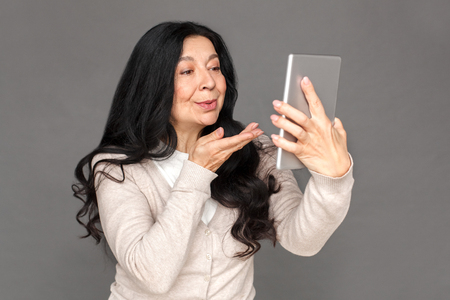 Freestyle. Mature lady standing isolated on grey sending kiss to camera having call on digital tablet close-upの写真素材