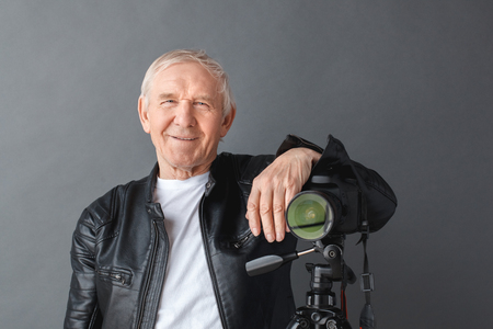 Senior man in leather jacket standing isolated on gray leaning on camera on tripod smiling playful close-upの写真素材
