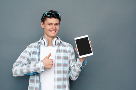 Man wearing headphones isolated on gray wall tourism concept standing holding digital tablet thumb up cheerfulの写真素材