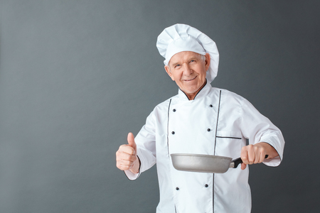 Senior chef studio standing isolated on gray holding pan looking camera cheerful thumb upの写真素材