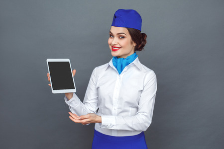 Professional Occupation. Stewardess standing isolated on grey showing screen of digital tablet smiling friendlyの写真素材
