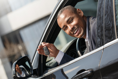 Young man sitting in new car looking out from window with key looking camera playfulの写真素材