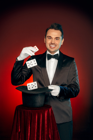 Professional Occupation. Magician in suit and gloves standing isolated on wall showing trick with cards and hat smiling cheerfulの写真素材