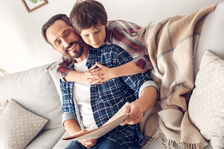 Father and little son at home boy hugging dad sitting on sofa reading newspaper cheerfulの写真素材