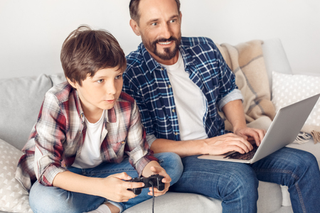 Father and little son at home sitting on sofa dad typing on laptop looking curious at boy playing game console excitedの写真素材