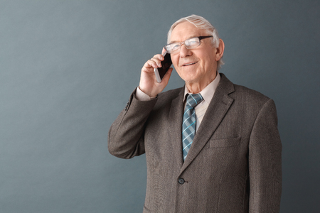 Senior man teacher wearing glasses studio standing isolated on gray answering phone call looking camera happyの写真素材