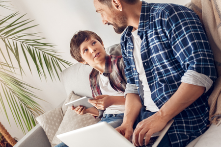 Father and little son at home sitting on sofa dad working on laptop while boy using digital tablet discussing pastimeの写真素材