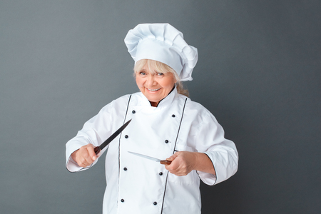 Senior woman chef studio standing isolated on gray with knives frightening playfulの写真素材