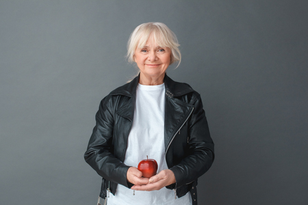 Senior woman in leather jacket studio standing isolated on gray holding apple looking camera happy calmの写真素材