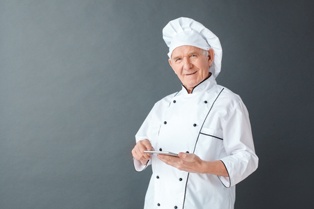 Senior chef studio standing isolated on gray with digital tablet looking camera cheerfulの写真素材