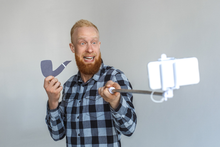 Freestyle. Mature man standing isolated on grey with monopod taking selfie on smartphone with tobacco pipe smiling cheerfulの写真素材