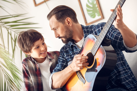 Father and little son at home sitting on sofa boy smiling looking at dad playing guitarの写真素材