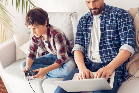 Father and little son at home sitting on sofa dad working on laptop concerned while boy playing game concentratedの写真素材