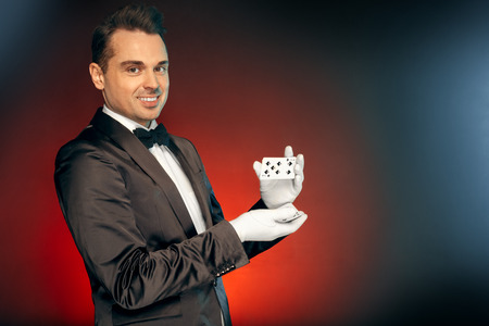 Professional Occupation. Magician in suit and gloves standing isolated on wall showing trick shuffling cards smiling playfulの写真素材