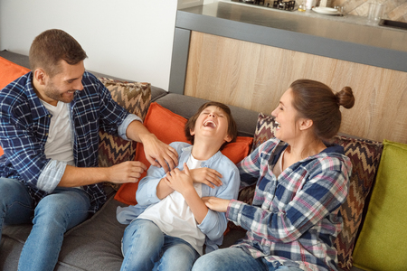Family at home sitting on couch in living room together parents tickling son playfulの写真素材