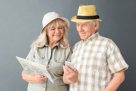 Senior tourists in beach hats studio standing isolated on gray with map woman showing husband pictures on smartphone happyの写真素材