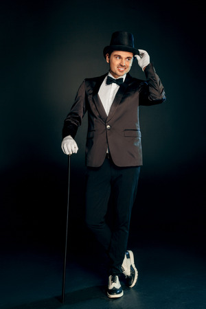Professional Occupation. Showman in suit gloves and hat standing isolated on wall with cane posing looking aside smiling playful full body shotの写真素材