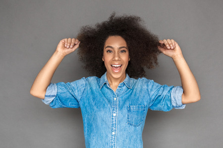 Freestyle. Mulatto woman standing isolated on grey holding hair looking camera playfulの写真素材