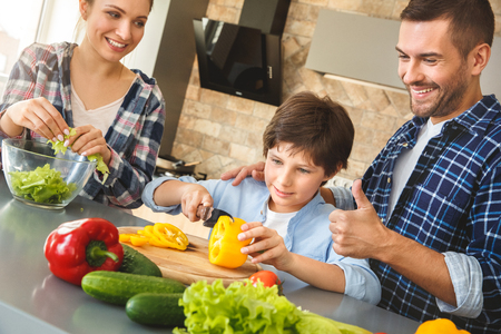 Family at home standing in kitchen together father hugging son cutting bell pepper showing thumb up smiling proudの写真素材