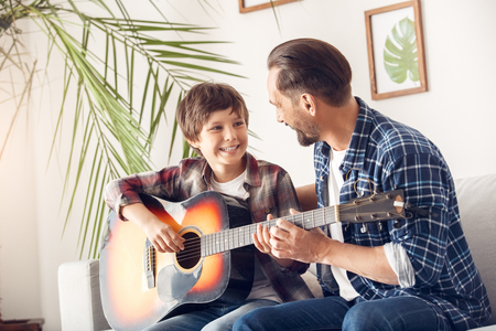 Father and little son at home sitting on sofa boy playing guitar looking at dad cheerfulの写真素材
