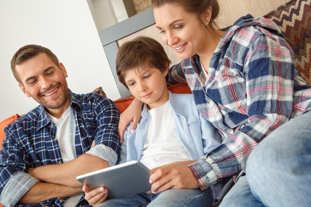Family at home sitting on couch in living room playing digital tablet together smiling excitedの写真素材