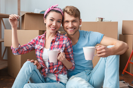 Young couple moving to new place sitting holding tea cups and keys looking camera joyfulの写真素材