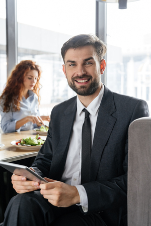 Businesspeople having business lunch at restaurant sitting man close-up holding digital tablet looking camera smiling while woman eating saladの写真素材