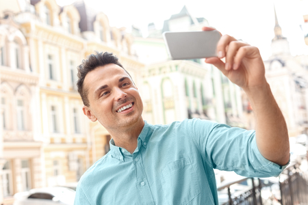 Capturing moments. Young man standing on city street taking selfie on smartphone looking camera cheerful close-upの写真素材