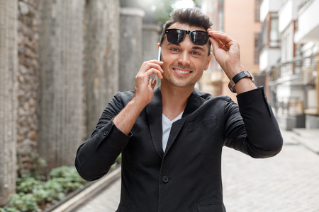 Outdoors leisure. Young man in suit and sunglasses on city street taking off sunglasses answering call looking camera playfulの写真素材