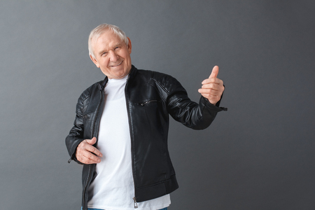 Senior man in leather jacket standing isolated on gray playing imaginary guitar looking camera cheerfulの写真素材