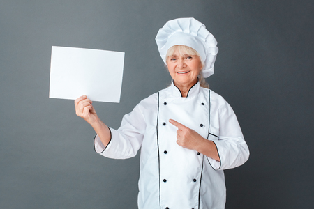 Senior woman chef studio standing isolated on gray pointing at paper looking camera happyの写真素材