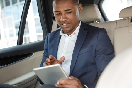 Young businessman sitting on back seat in car playing game on digital tablet joyfulの写真素材