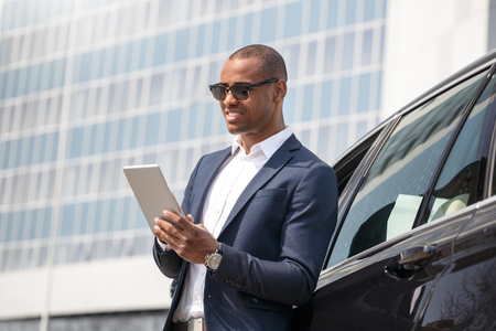 Young man in sunglasses leaning on car browsing digital tablet smiling cheerfulの写真素材
