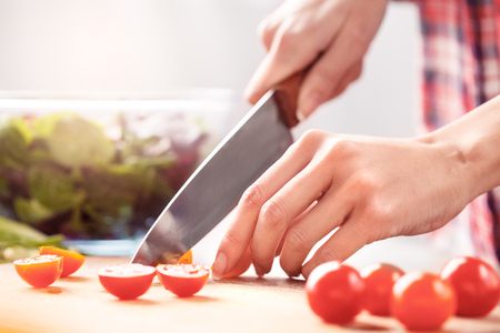 Young girl at kitchen healthy lifestyle standing holding knife cutting tomatoes close-upの写真素材