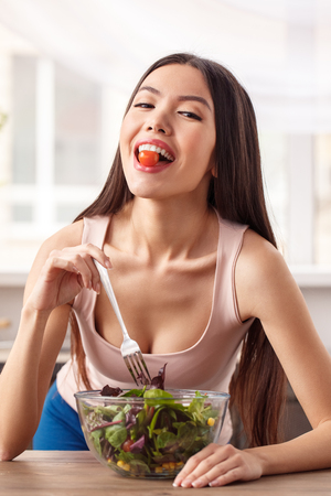 Young girl at kitchen healthy lifestyle leaning on table eating salad holding tomato with teeth looking camera cheerfulの写真素材
