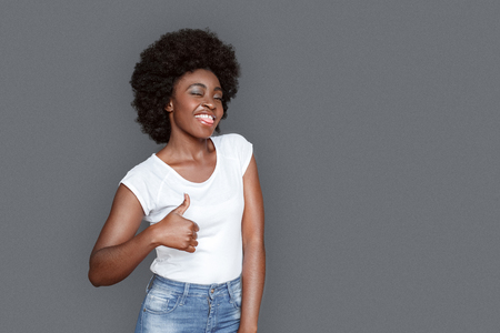 Young woman standing isolated on gray thumb up tongue out looking camera playfulの写真素材