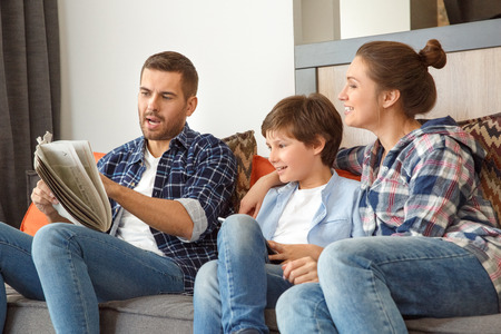 Family at home sitting on couch in living room together father showing newspaper shocked to mother and son holding digital tablet happyの写真素材