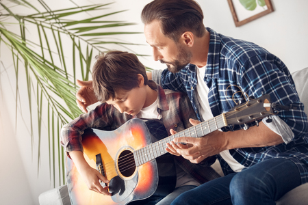 Father and little son at home sitting on sofa dad teaching boy how to play guitarの写真素材