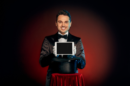 Professional Occupation. Magician in suit and gloves standing isolated on wall making trick taking digital tablet from hat on table smiling friendlyの写真素材