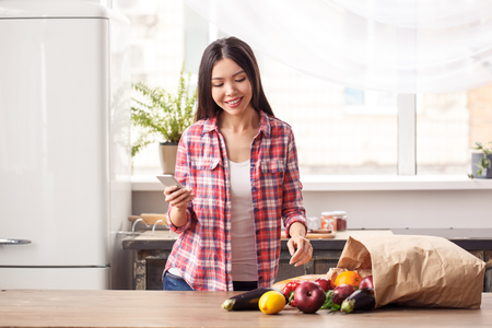 Young girl at kitchen healthy lifestyle standing holding hpone looking at products bag on table cheerfulの写真素材
