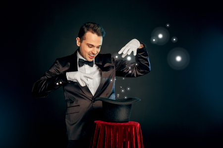 Professional Occupation. Magician in suit and gloves standing isolated on wall making trick with hat on table smiling playfulの写真素材