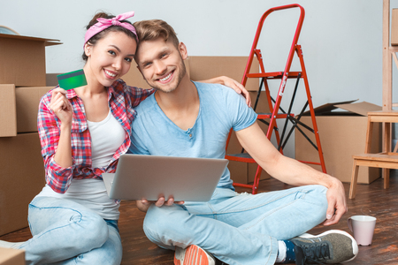 Young couple moving to new place sitting with laptop holding credit card smiling cheerfulの写真素材