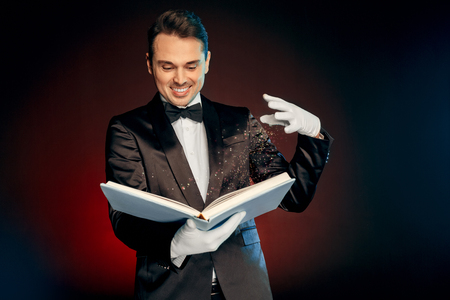 Professional Occupation. Magician in suit and gloves standing isolated on wall making spell with magic book smiling excitedの写真素材