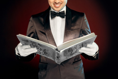 Professional Occupation. Magician in suit and gloves standing isolated on wall reading magic book joyful close-upの写真素材